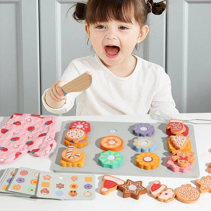 Child holding a toy knife beside a tray of wooden cookies, pattern cards, and a pink oven mitt.