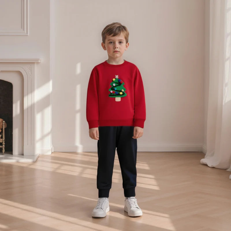 Child wearing a red Christmas tree sweatshirt with black pants and white sneakers near a fireplace.