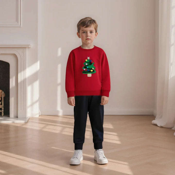 Child wearing a red Christmas tree sweatshirt with black pants and white sneakers near a fireplace.