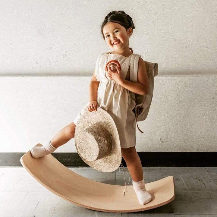 Child balancing on a curved wooden board while holding a straw hat