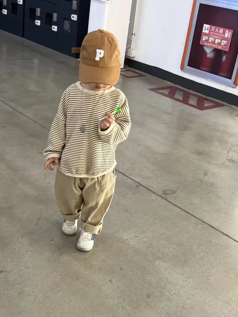 Child wearing khaki pants with a striped top, brown cap, and white sneakers