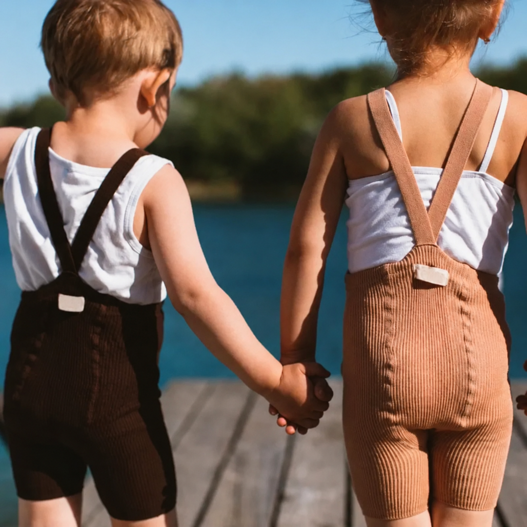 Two children wearing black and brown suspender shorts with crossed straps over white tops.