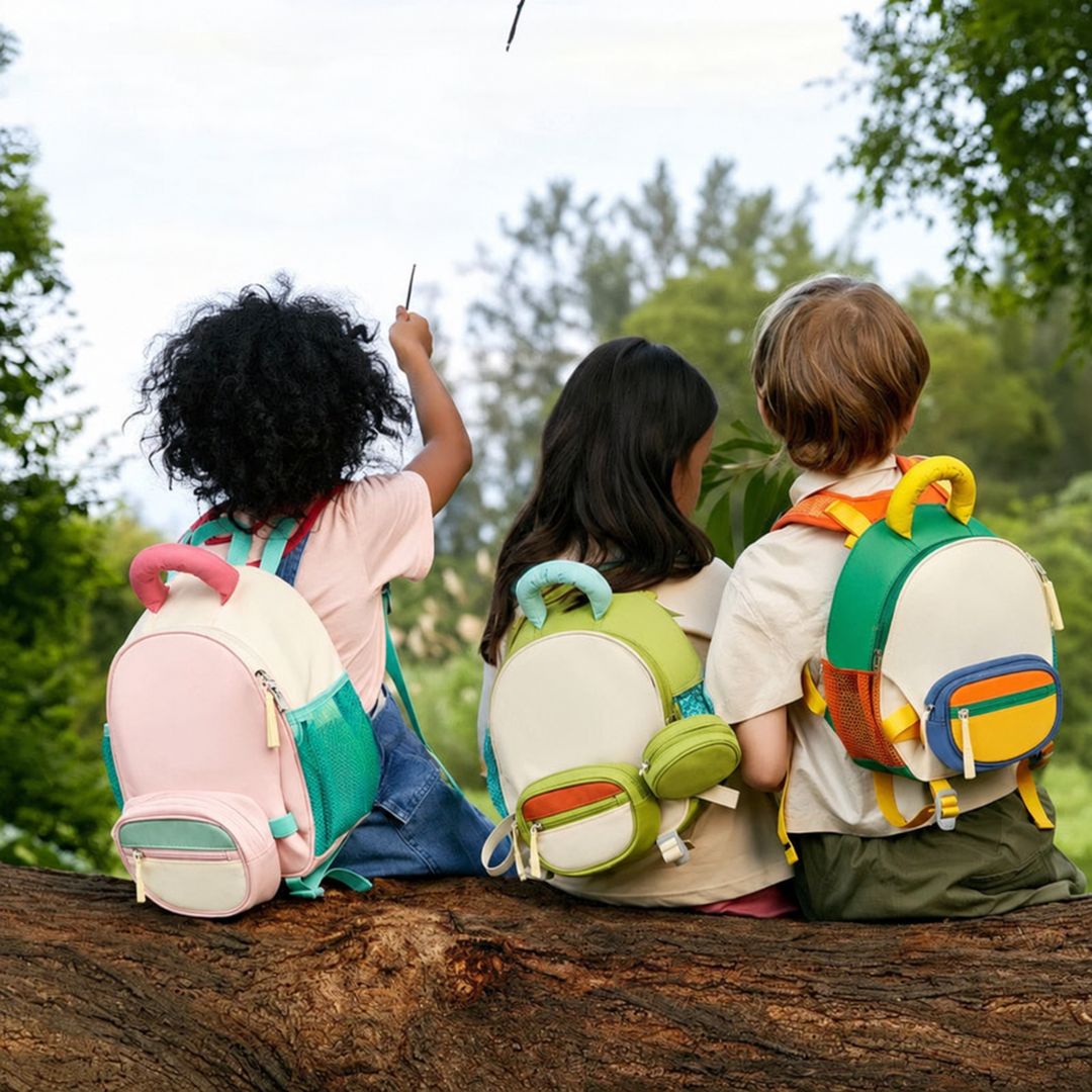 Three children wearing colorful backpacks while sitting on a fallen tree outdoors