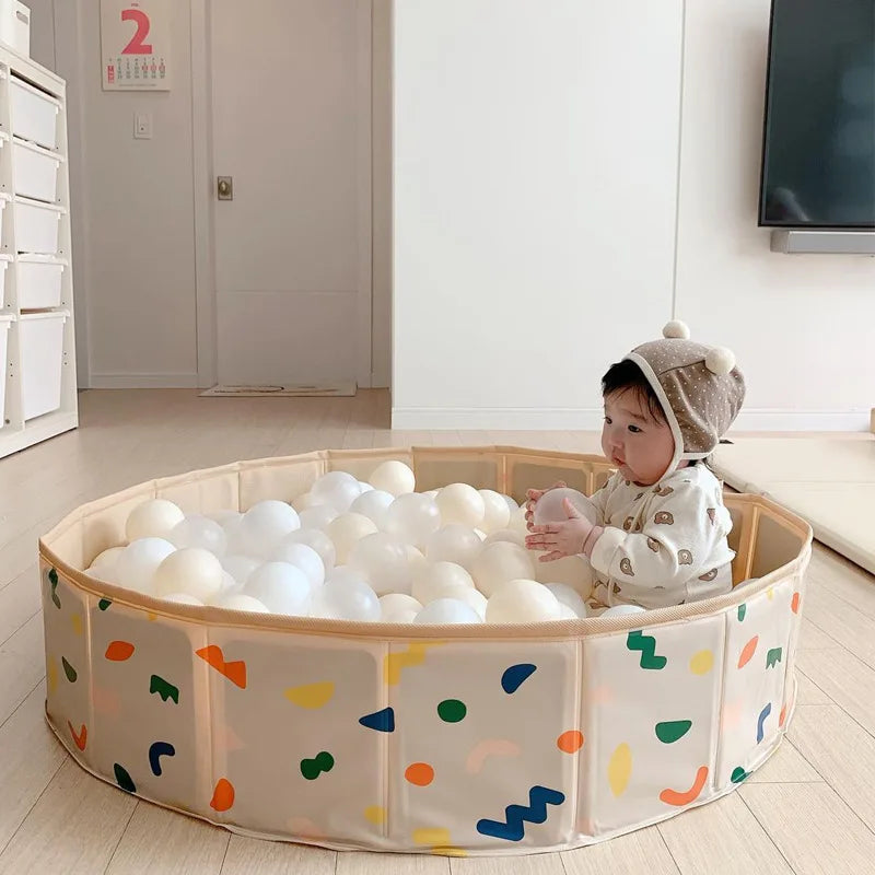 Baby sitting among white play balls in a round, multicolored play pool indoors