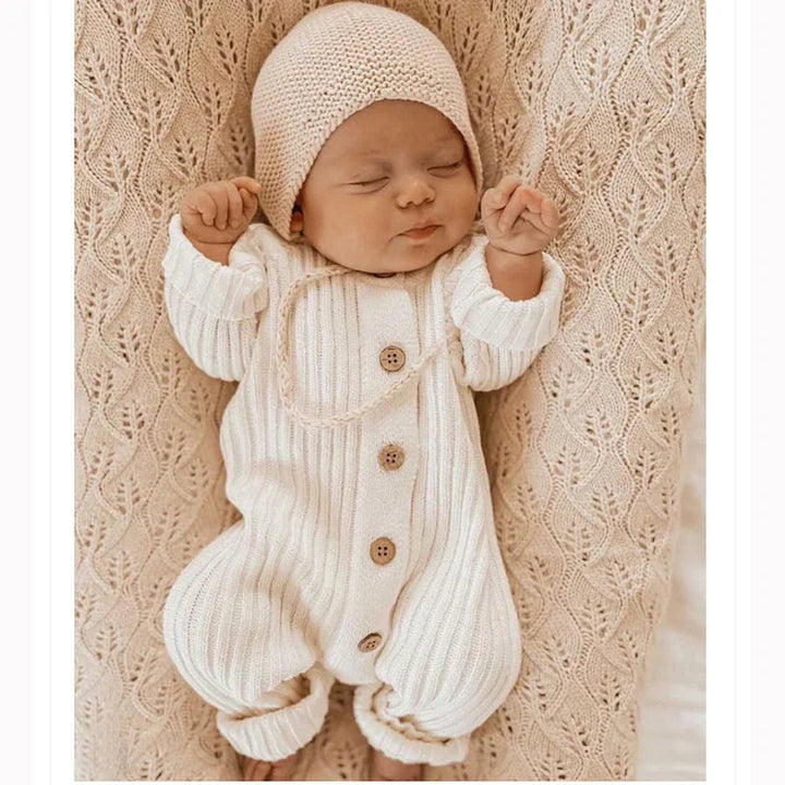 Baby wearing a cotton-knit romper and beige hat on a textured blanket