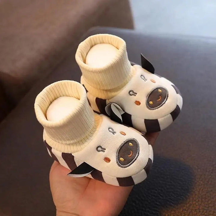 Cream cow indoor shoes held in one hand, with ribbed cuffs and black-and-white details