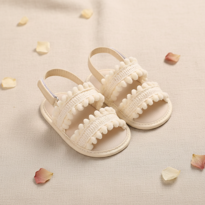 Cream open-toe baby sandals with pom-pom trim displayed beside scattered flower petals.