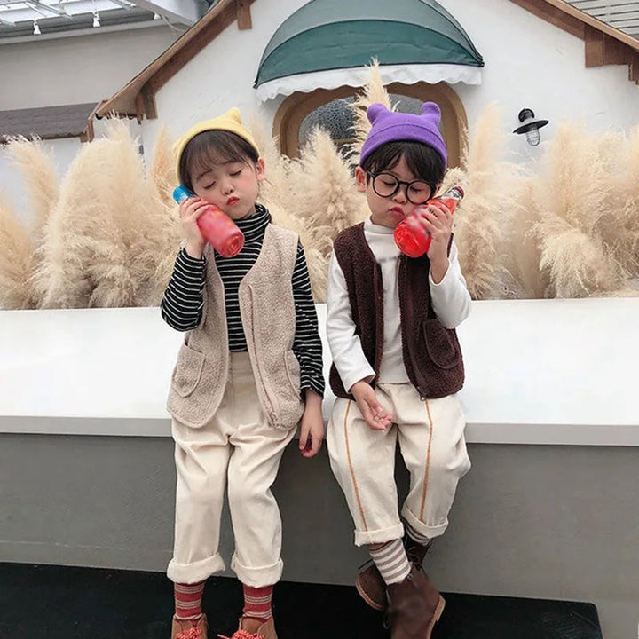 Two children wearing light-colored and brown fleece vests with layered outfits.