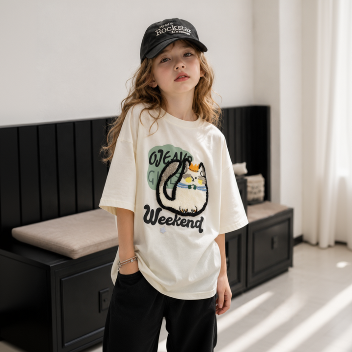 Child standing in a cream oversized cat-print tee with black shorts and a cap