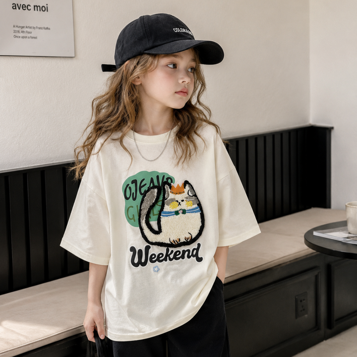 Child wearing a cream cat-print T-shirt with black shorts and a black cap