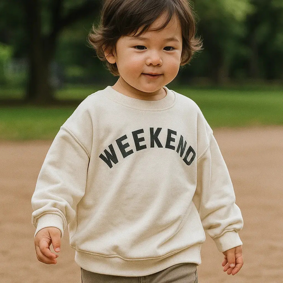 Child outdoors wearing a cream crewneck sweater with dark Weekend lettering.