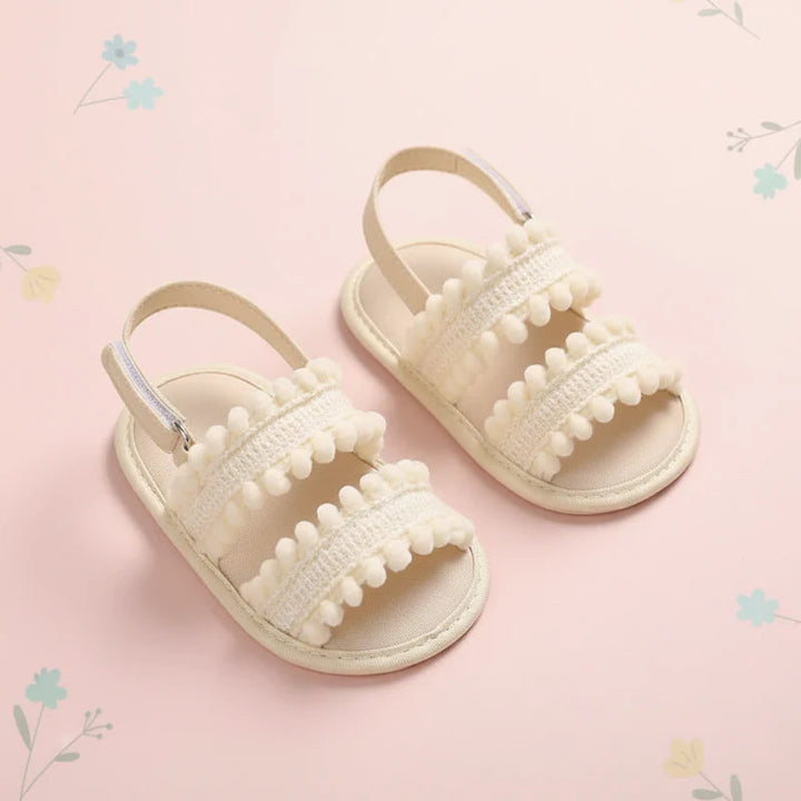 Cream baby sandals with pom-pom trim and heel straps on a pink floral background.