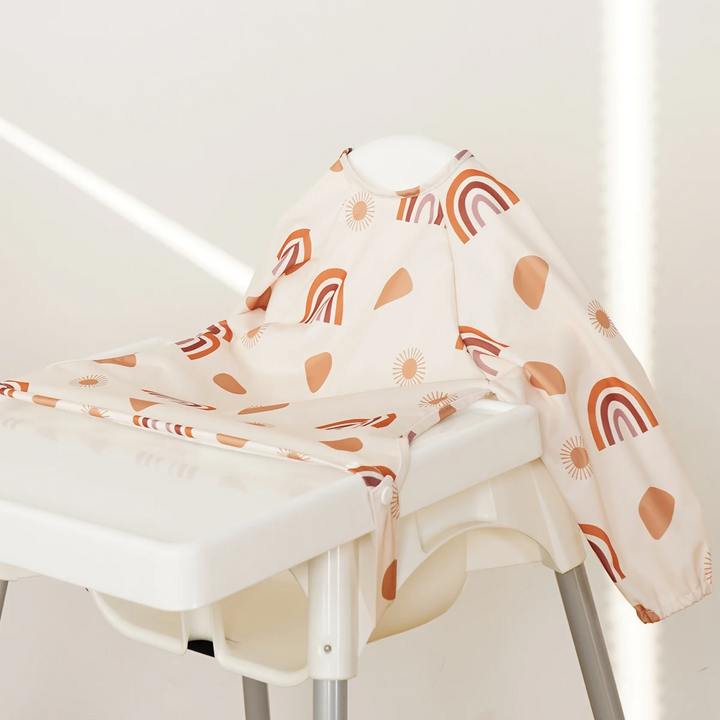 Cream long-sleeve baby bib with orange rainbows spread over a high-chair tray