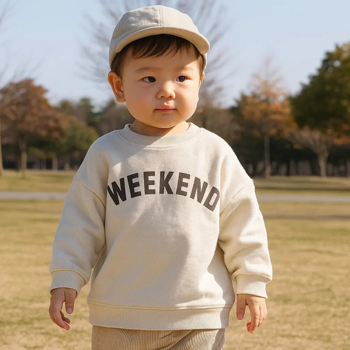 Child in a cream Weekend sweater and beige cap standing on grass.