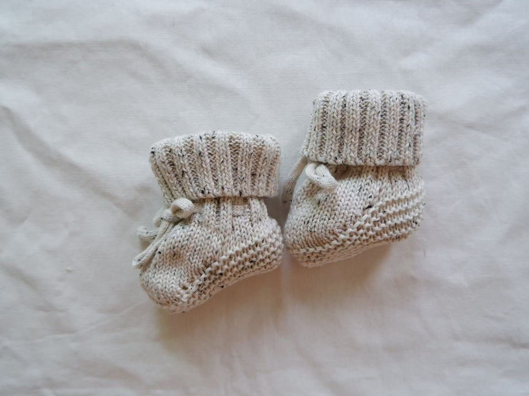 Light knitted baby booties with dark flecks, folded cuffs, and ankle ties.