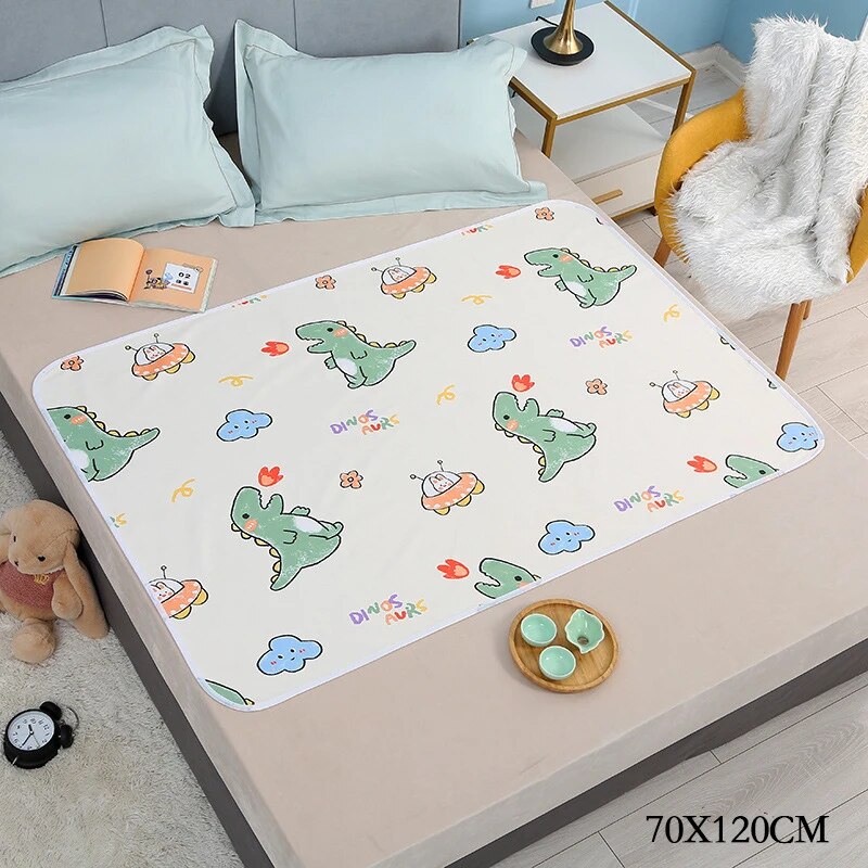White changing mat with green dinosaurs and blue clouds on a bed, labeled 70 x 120 cm.