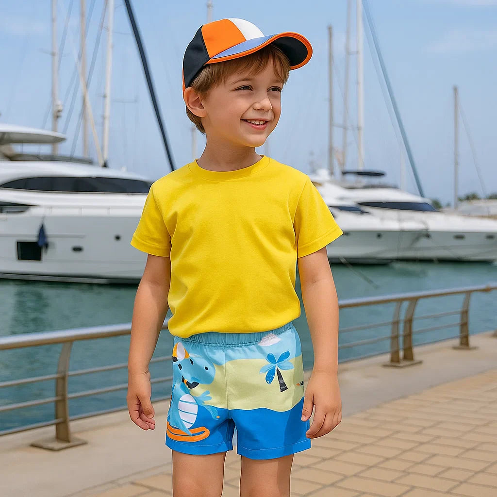 Child wearing blue dinosaur-print swim trunks and a yellow T-shirt beside a marina.