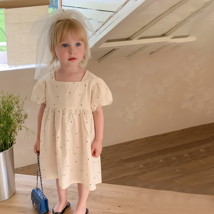 Full-length front view of the dotted A-line dress worn by a child beside a wooden table.