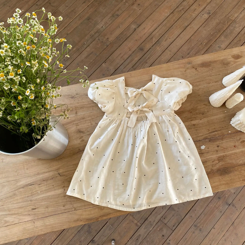 White dotted dress laid on a wooden table beside flowers, with both back bows visible.
