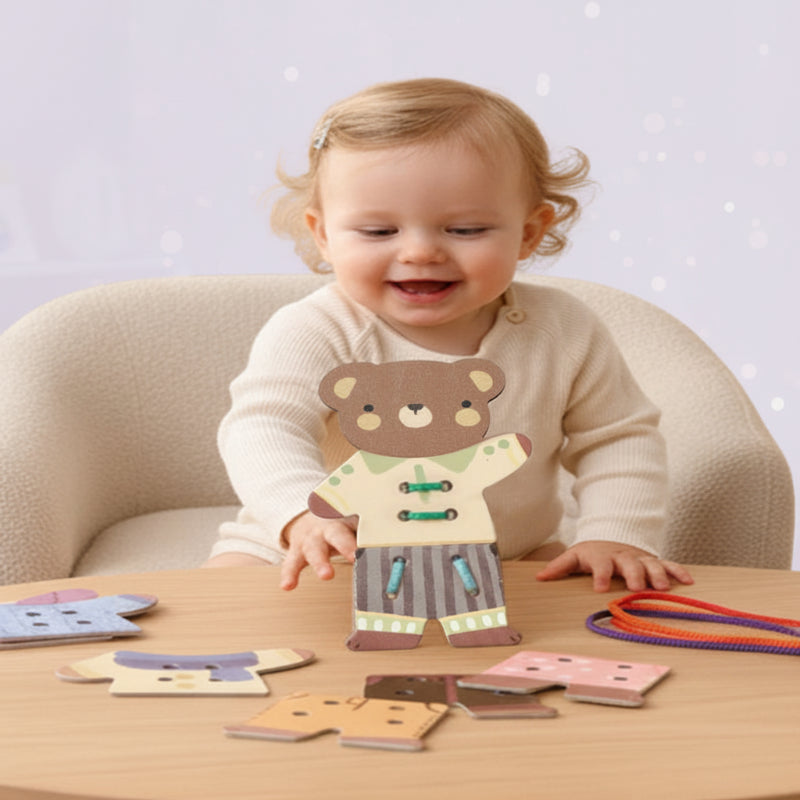 A child holding a dressed wooden bear at a table with clothing pieces and cords.