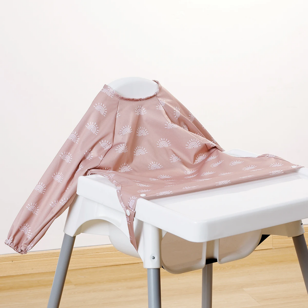 Dusty pink long-sleeve baby bib with white sunburst motifs on a high chair