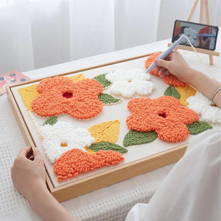 Hands using a punch needle on a framed orange and white floral embroidery design.