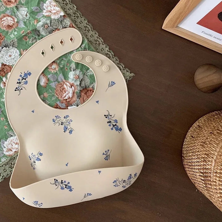 Beige silicone baby bib with blue floral print and a wide food-catching pocket