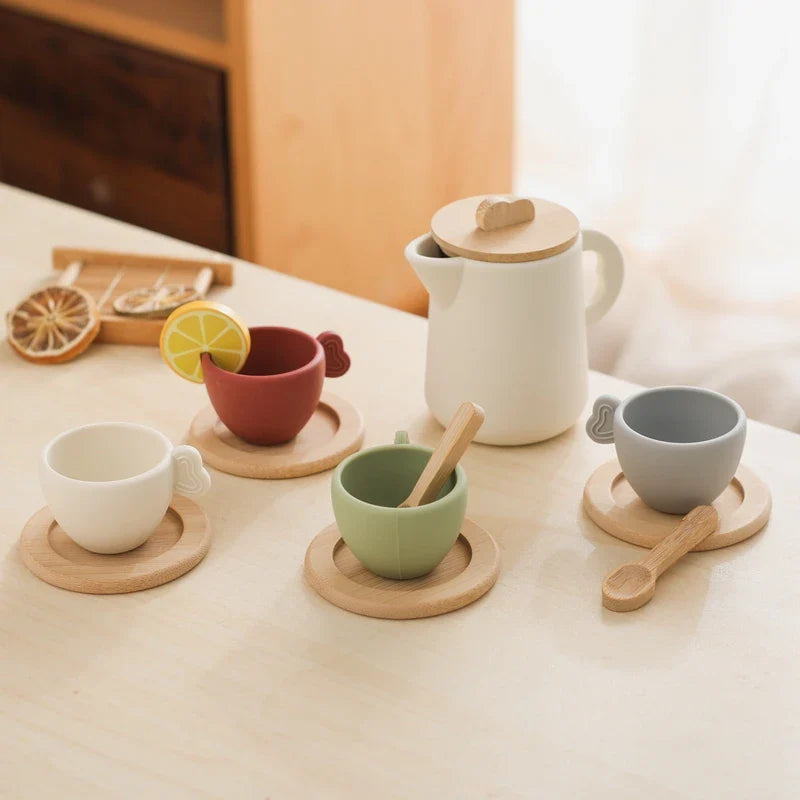 Four colored tea cups on wooden saucers beside a lidded pitcher and play spoons