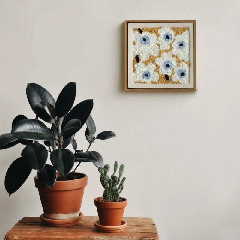 Framed white flower embroidery hanging on a wall above potted plants.