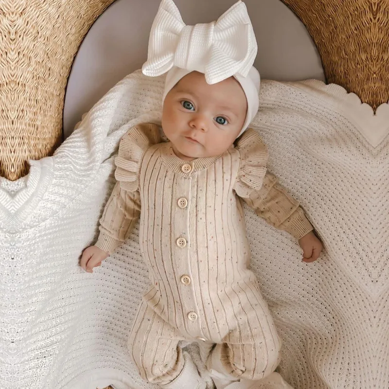 Full-length view of a baby wearing a beige knitted romper and white bow headband