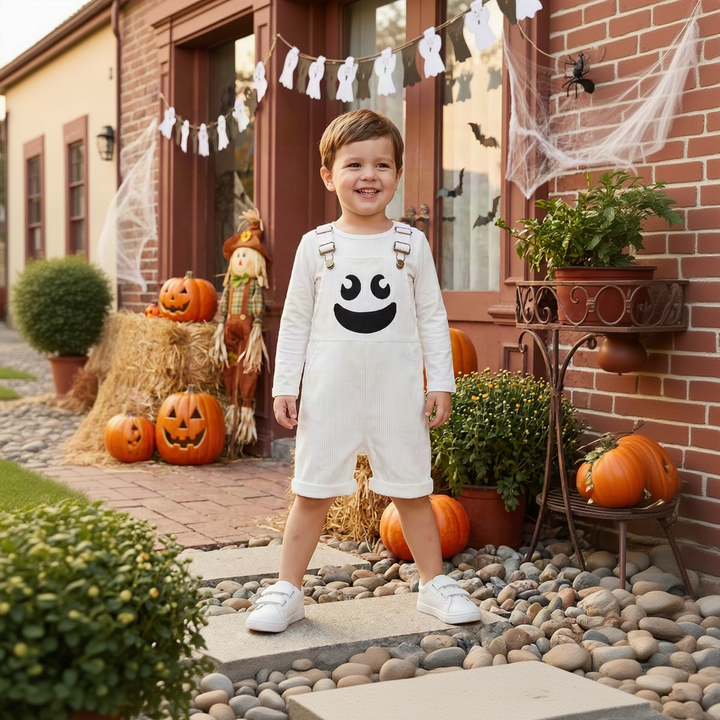 Child wearing white ghost overalls over a long-sleeve top beside pumpkins and a ghost garland.