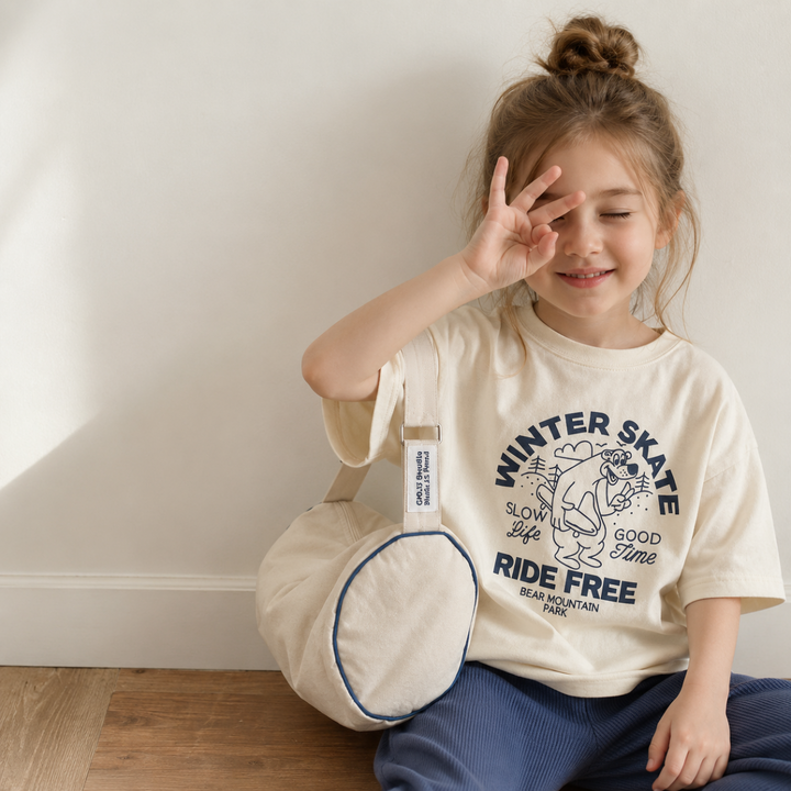 Girl wearing a cream skate bear T-shirt with blue pants and a round shoulder bag