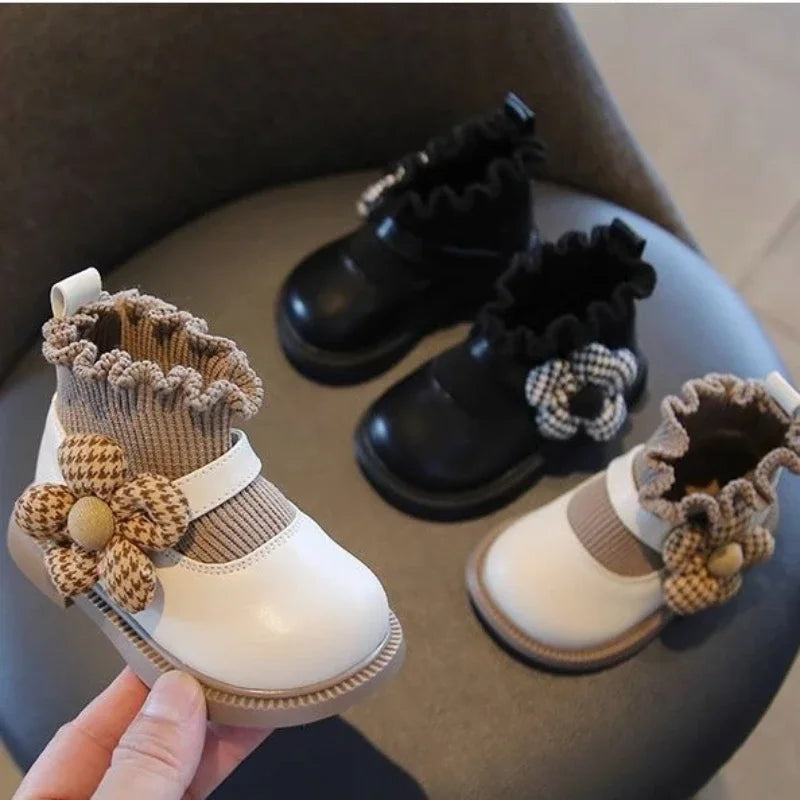 White flower-accented girls' boot held in front of white and black pairs on a chair