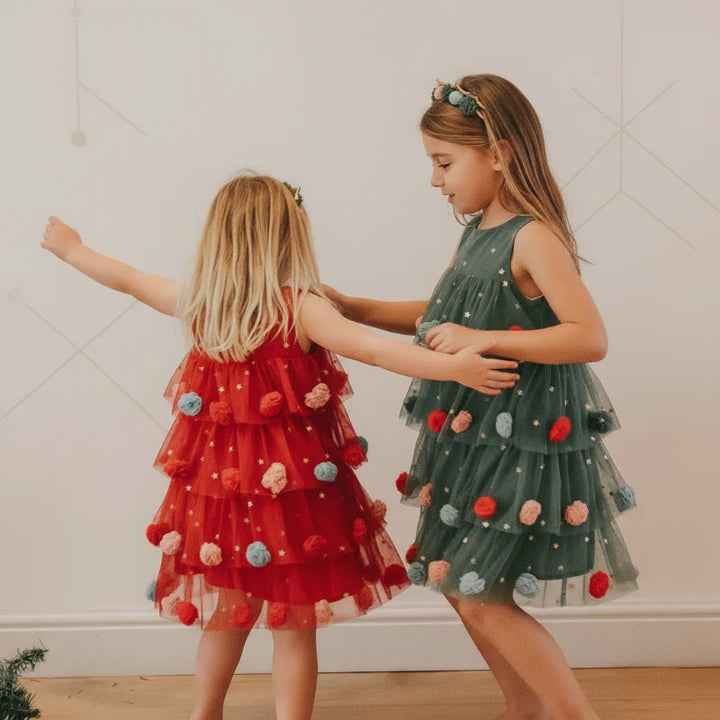 Two girls wearing red and green Christmas dresses with tiered skirts and colorful pom-pom details