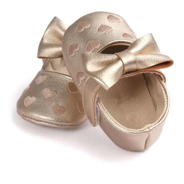 Overlapping gold baby shoes showing bow accents, heart details, and the interior