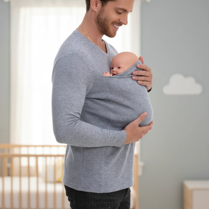 Man wearing a gray baby carrier shirt with a baby in the front pouch beside a crib.
