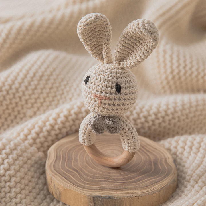 Gray crocheted bunny rattle standing on a wood slice beside a knitted blanket