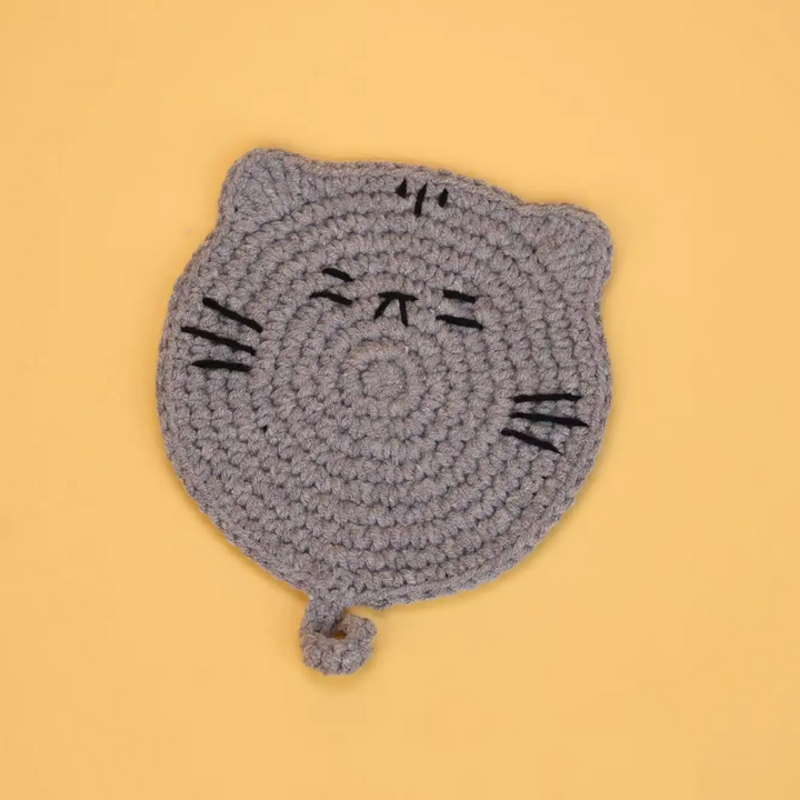Gray crocheted cat coaster with black stitched facial details and a curled tail