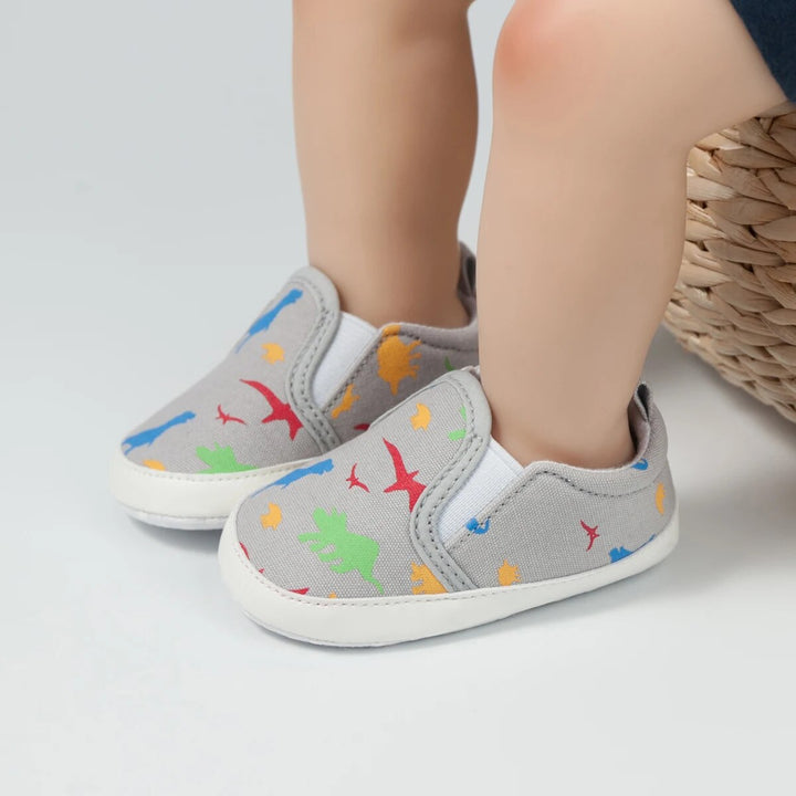 Gray dinosaur-print baby sneakers with white soles, shown on feet