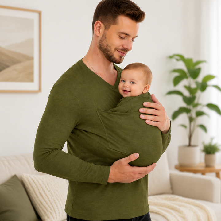 Man wearing a green long-sleeve baby carrier shirt with a baby in the front pouch in a living room.