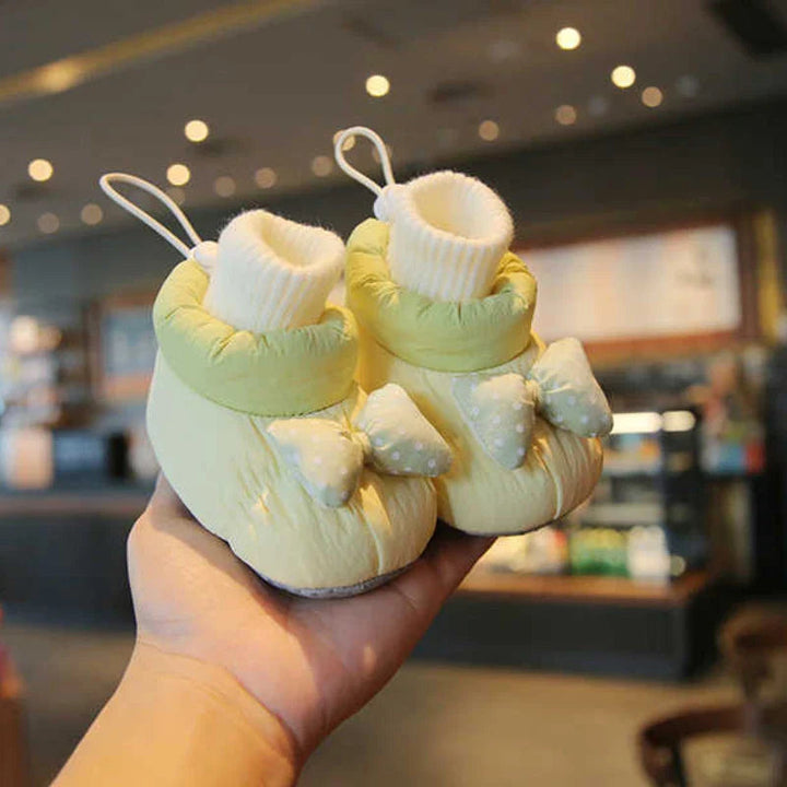 Green baby shoes with cream ribbed cuffs and bow details held in one hand
