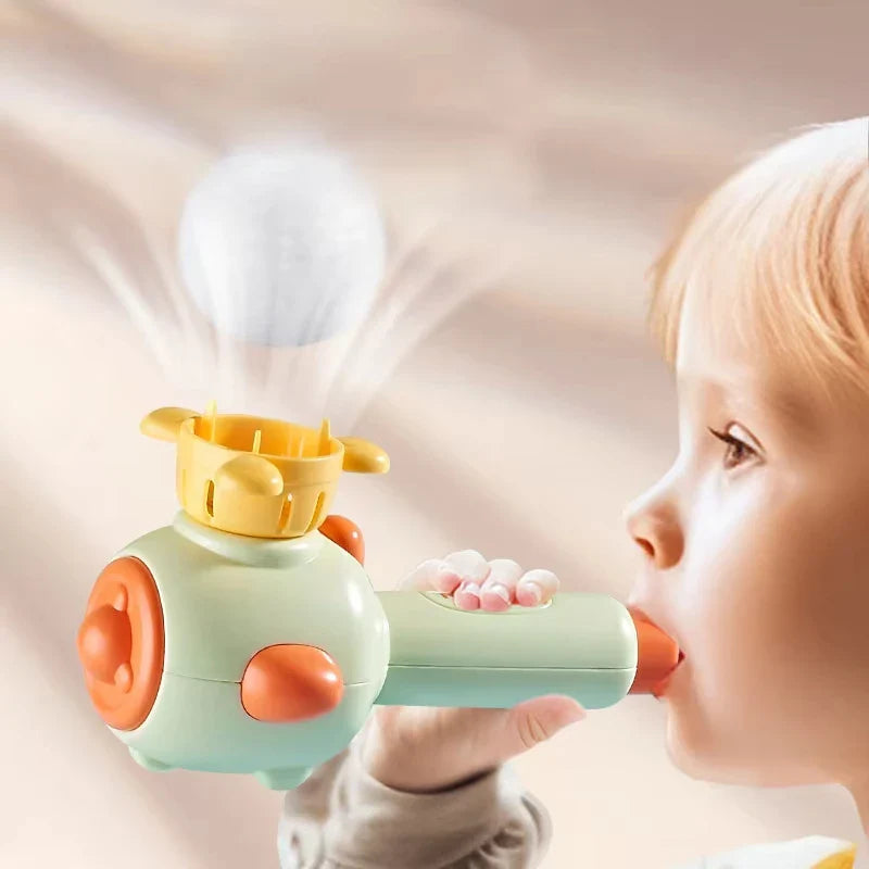 Child blowing into a green toy to lift a white ball above its yellow basket.