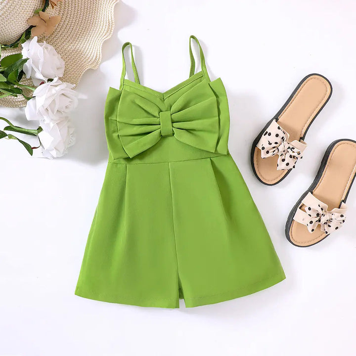Green bow-front romper laid flat beside sandals, white flowers and a straw hat