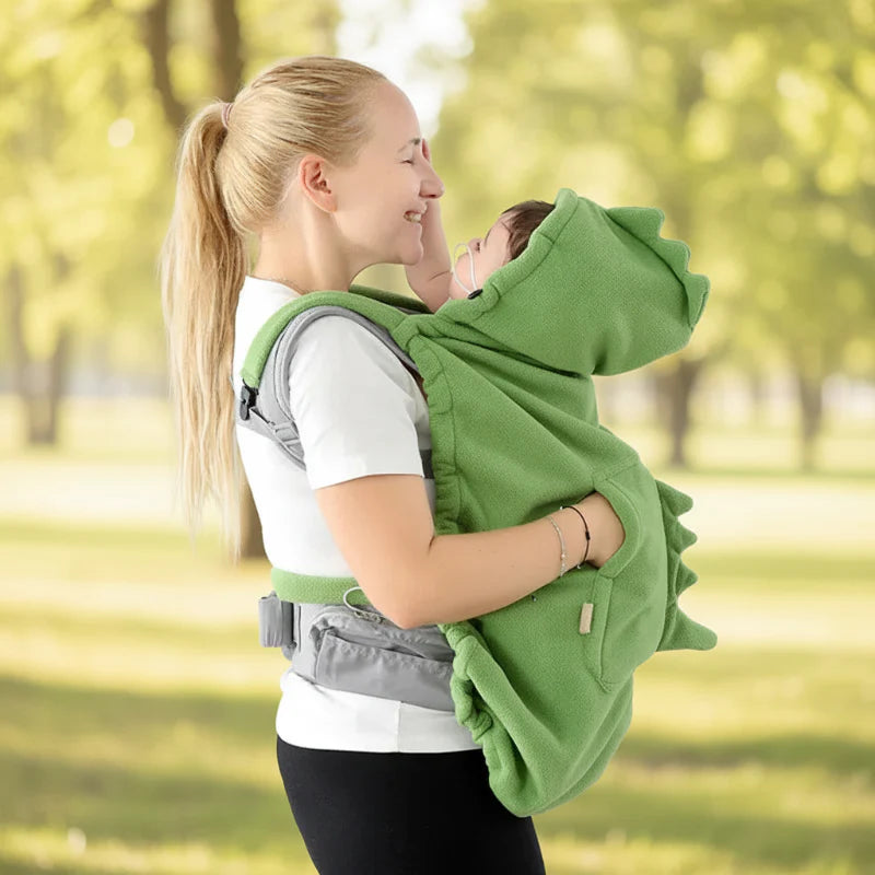 Parent wearing a green dinosaur baby carrier cover outdoors with the hood raised