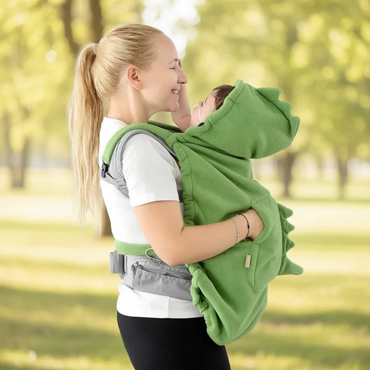 Parent wearing a green dinosaur baby carrier cover outdoors with the hood raised
