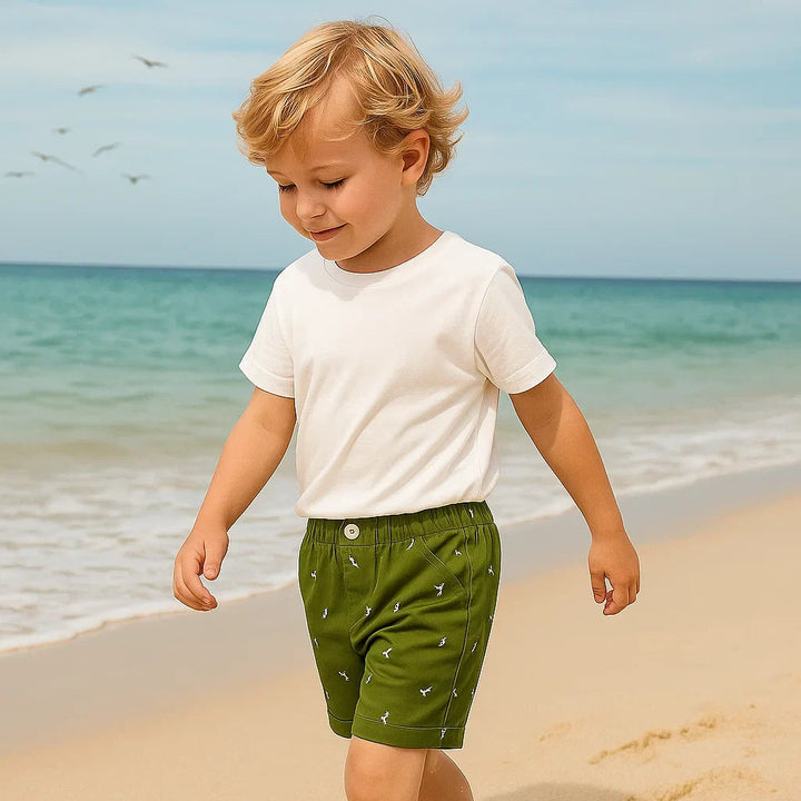 Child wearing forest green dinosaur shorts with a white T-shirt at the beach