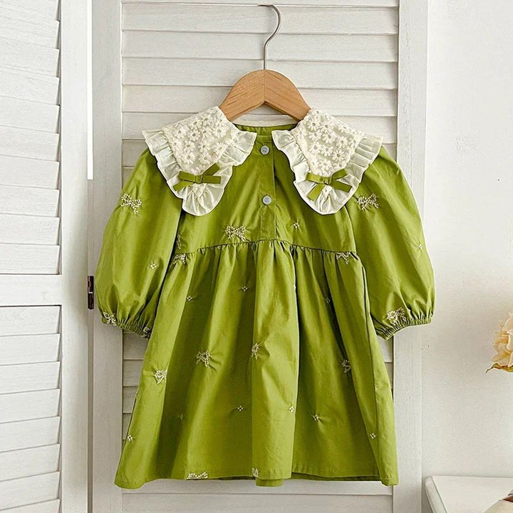 Green bow-embroidered dress with a gathered skirt hanging against white shutters