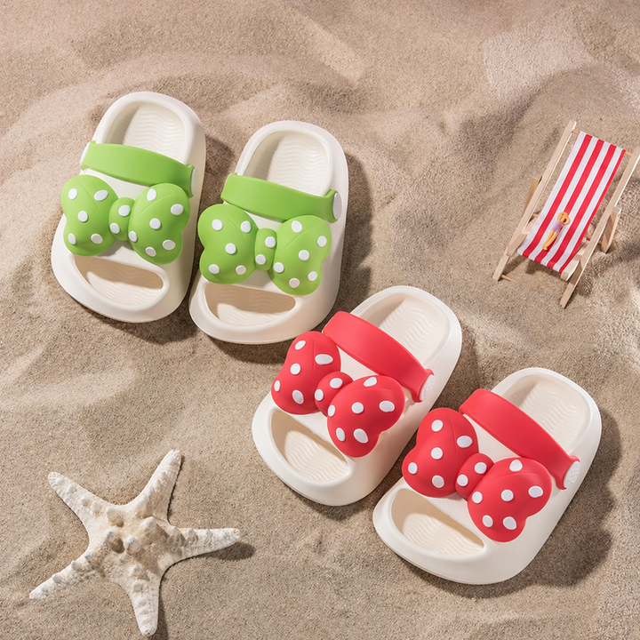 Green and red polka-dot bow sandals arranged on sand beside a starfish