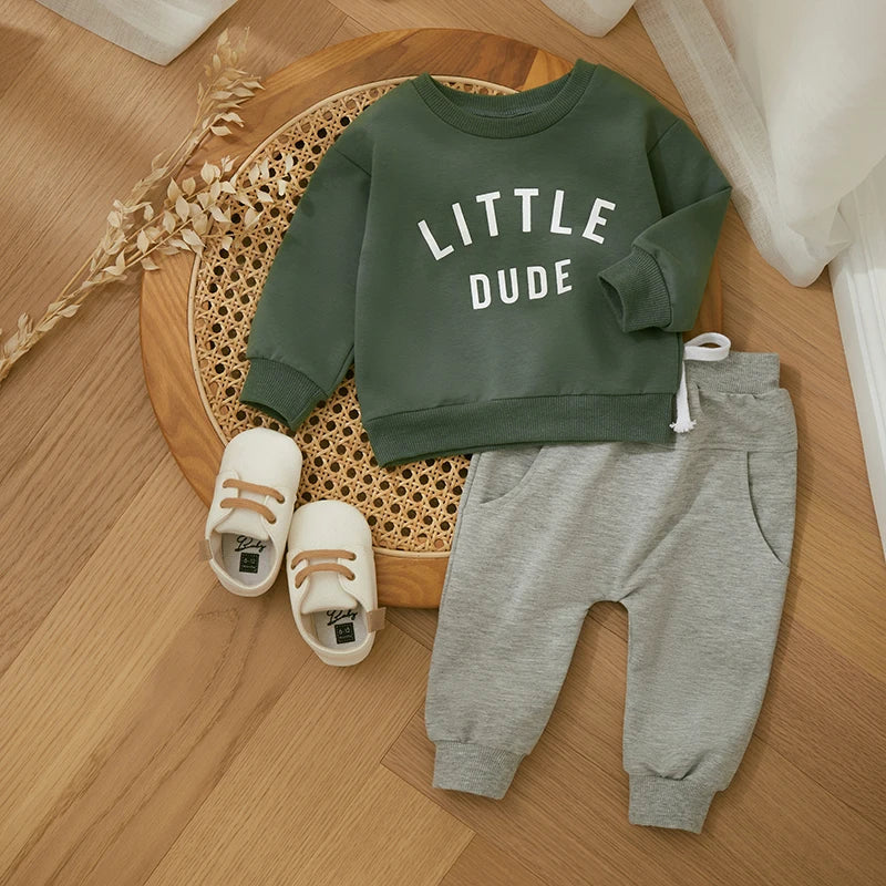 Green Little Dude sweatshirt and gray pants arranged with cream shoes on a woven chair
