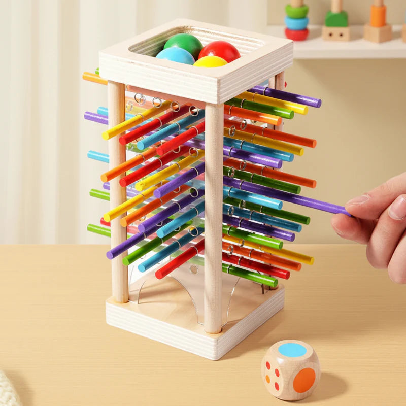 A hand pulling a purple stick from a wooden tower while colored balls rest at the top.